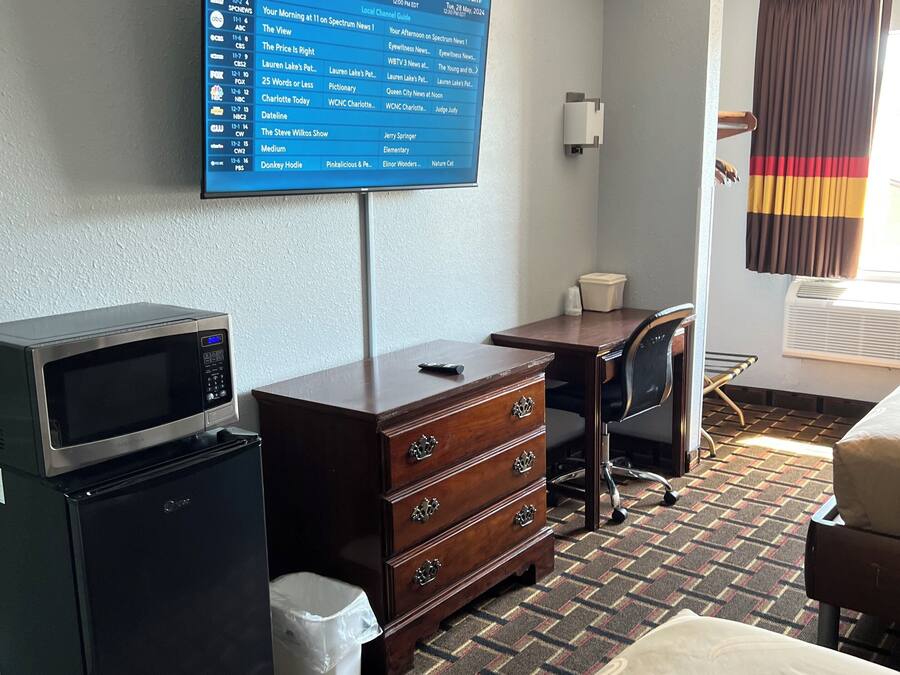 A hotel room with a television displaying a channel guide, a microwave, a refrigerator, a wooden dresser, a desk, a chair, and a bed.