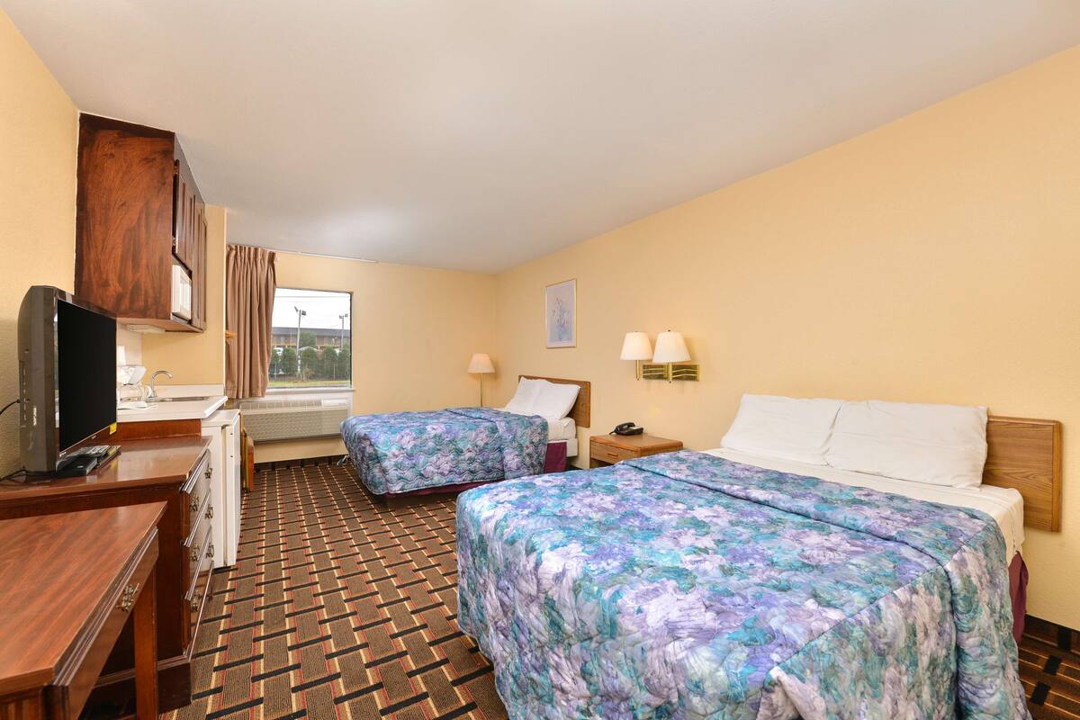 A hotel room with two beds, a desk with a TV, a window with curtains, and a patterned carpet.