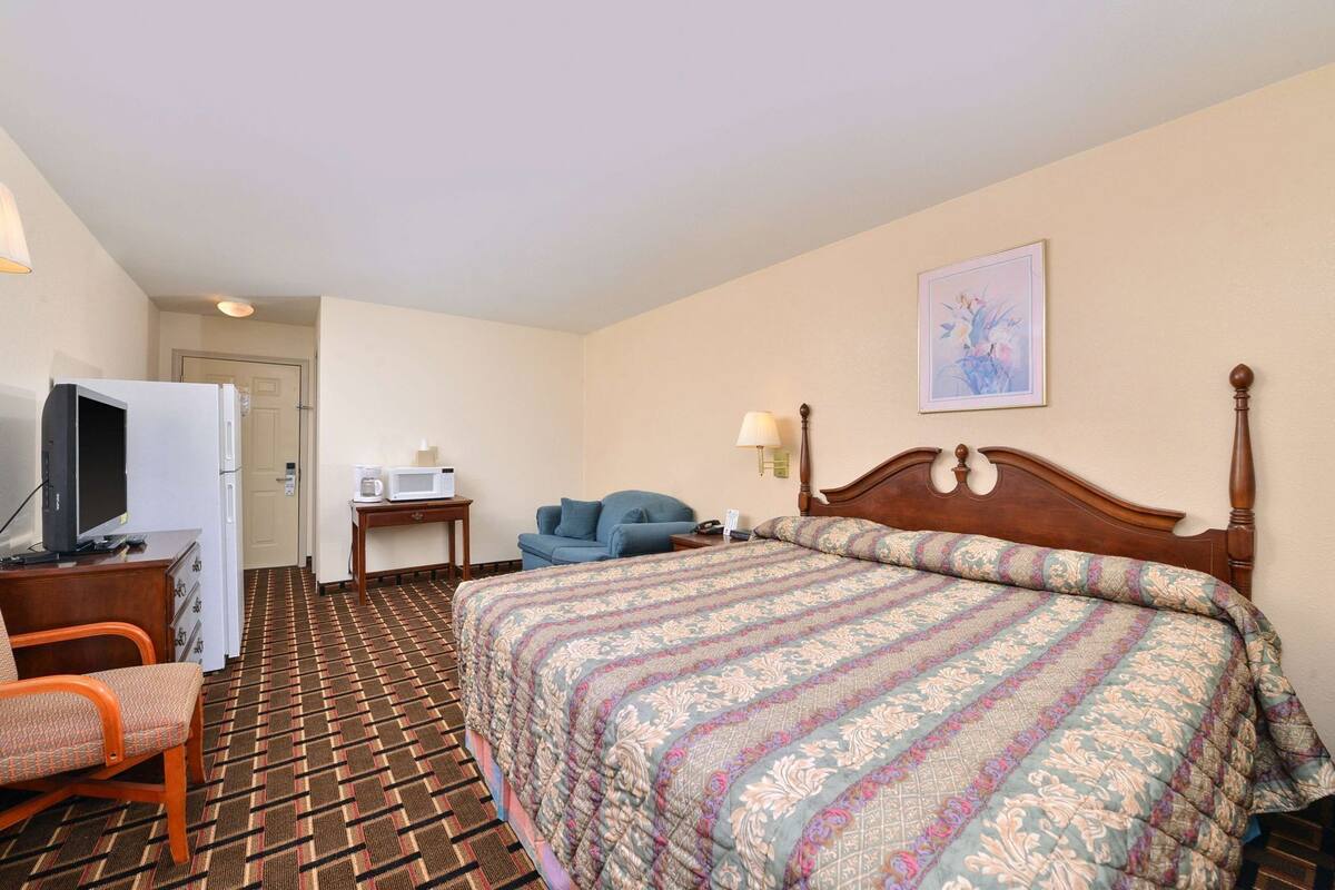 A hotel room with a bed, a desk with a television, a microwave, a chair, and a nightstand with a lamp.