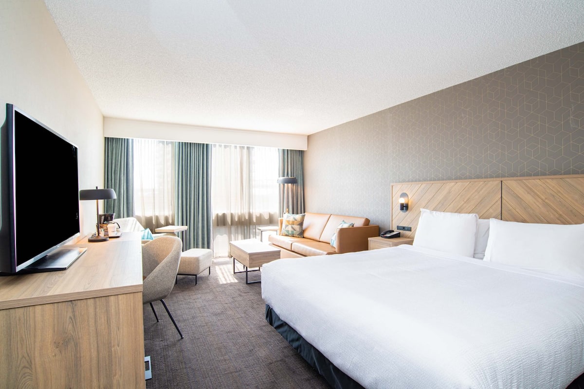 A hotel room with a large bed, a flat-screen TV, a desk with a lamp, a sofa, and a window with curtains.