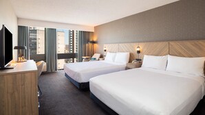A hotel room with two beds, a desk, a TV, and a view of the city.