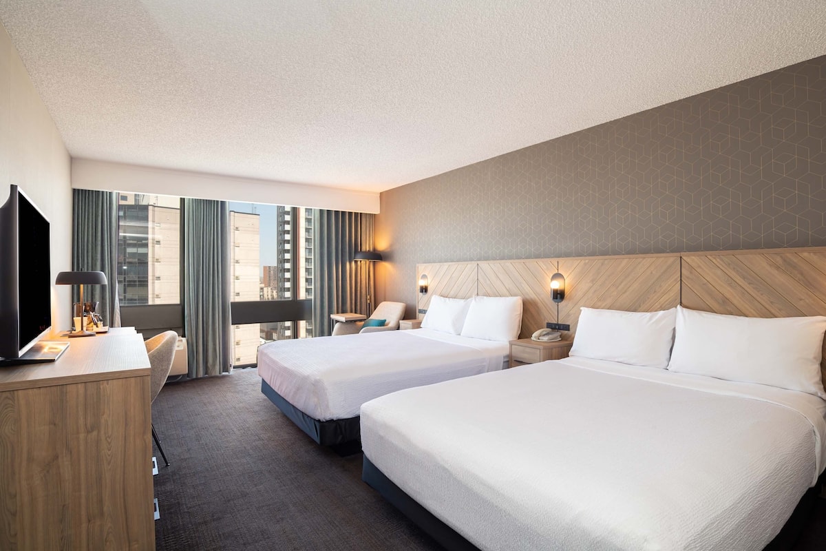 A hotel room with two beds, a desk, a TV, and a view of the city.