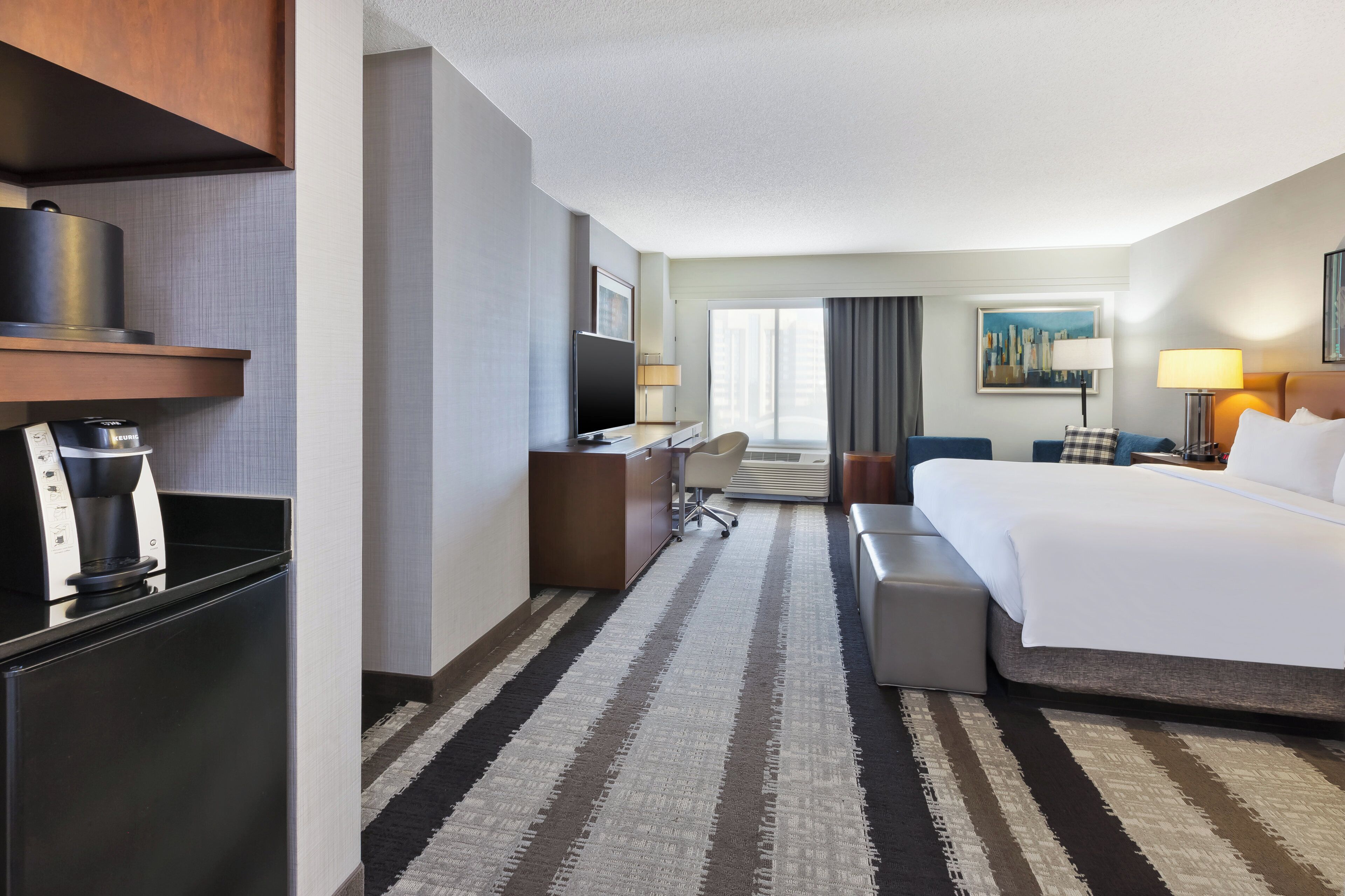 A hotel room with a bed, a desk, a chair, a coffee machine, and a large window.