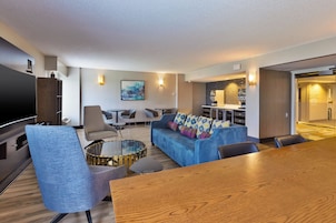 Quarto de hotel moderno com área de estar, incluindo um sofá azul, uma mesa de jantar de madeira e uma mesa de centro de vidro. Há uma televisão de tela plana na parede, uma pequena cozinha e uma porta que dá para outro cômodo.
