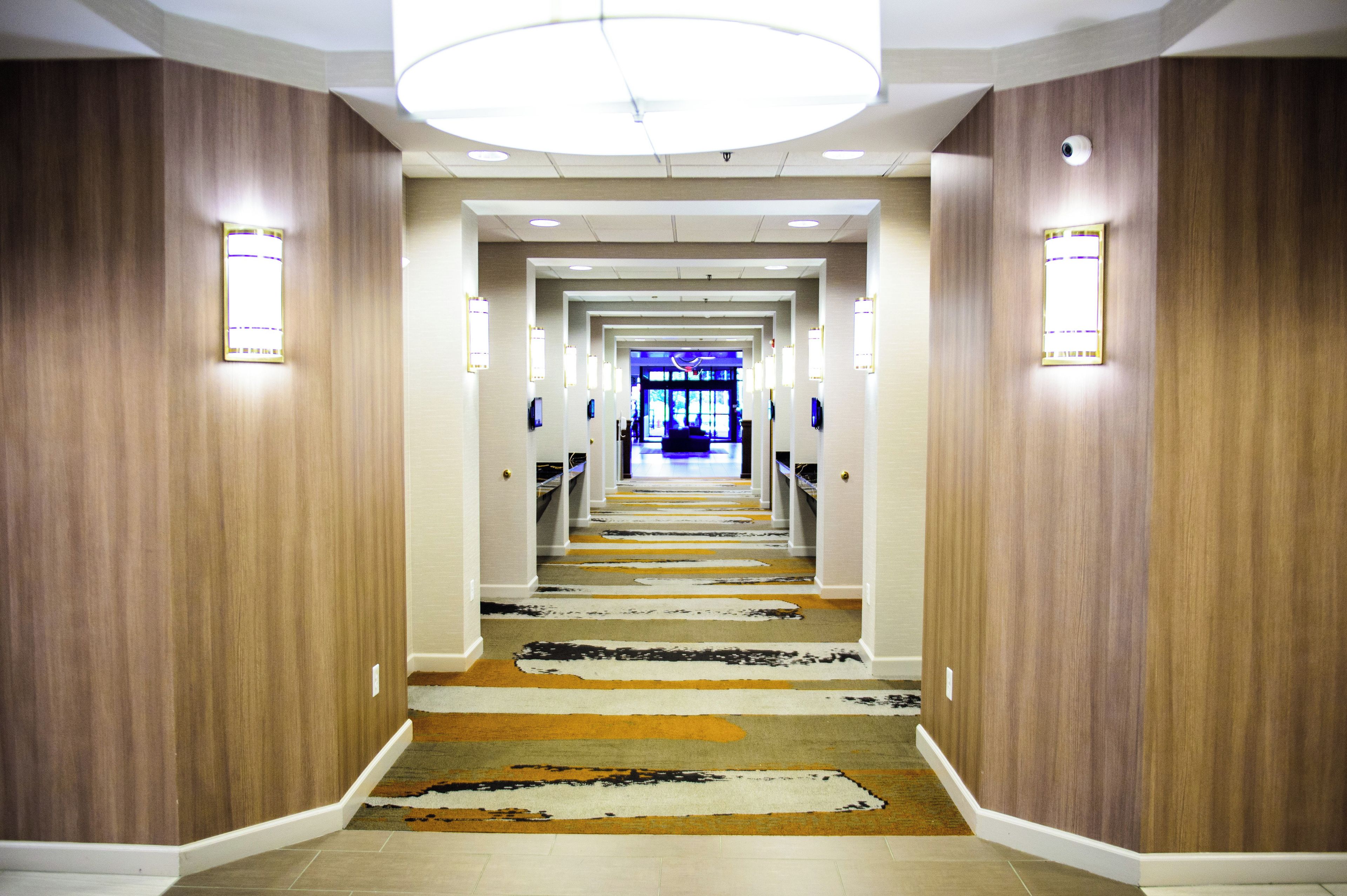 Un couloir d’hôtel aux murs lambrissés, recouvert d’un tapis à motifs, et une chambre illuminée de lumière bleue au bout du couloir.
