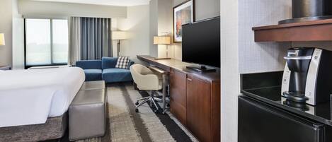A hotel room with a bed, a sofa, a desk with a TV, a coffee machine, and a large window.