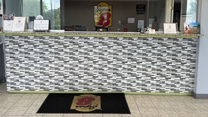 Un mostrador de recepción con un mostrador de registro, una pared con azulejos, un felpudo negro con un logotipo y una ventana con vistas a árboles.