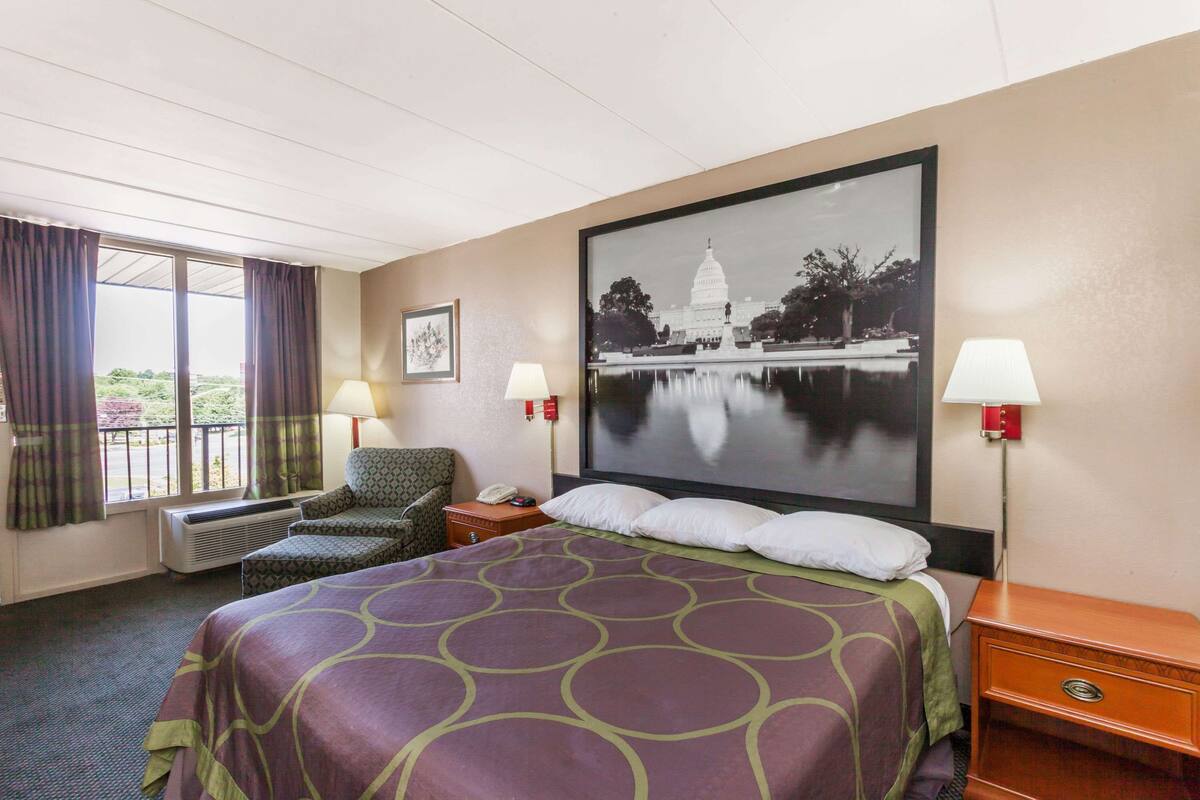 Ein Hotelzimmer mit einem großen Bett, zwei Nachttischlampen, einem Sessel, einem Schreibtisch mit Telefon und einem großen gerahmten Bild des Capitol-Gebäudes.