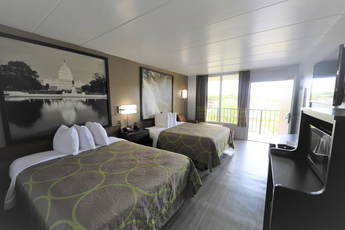 Ein Hotelzimmer mit zwei Betten, einem Fernseher, einem Schreibtisch und einem Balkon mit Blick auf ein Gebäude.