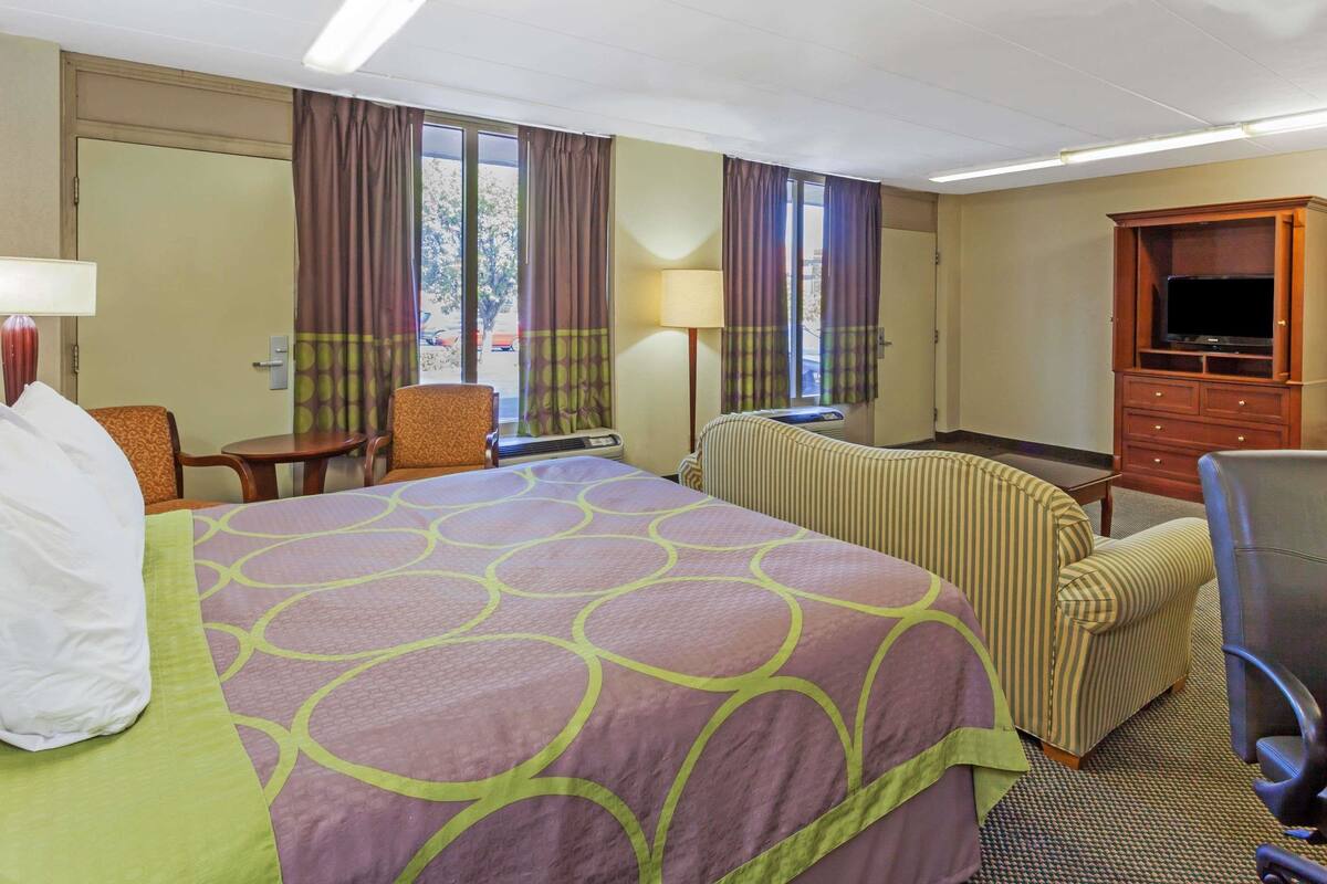 Ein Hotelzimmer mit einem großen Bett, einem Sessel, einem Schreibtisch, einem Fernseher und einem Fenster mit Vorhängen.