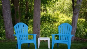 Dos sillas Adirondack azules y una pequeña mesa blanca en un área cubierta de césped, rodeada de árboles.