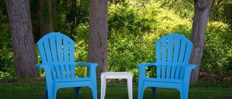 Deux chaises Adirondack bleues et une petite table blanche, installées sur une pelouse entourée d’arbres.