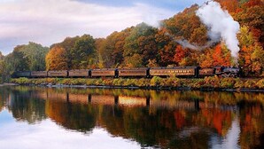 Un tren viaja a lo largo de un río con follaje otoñal al fondo.