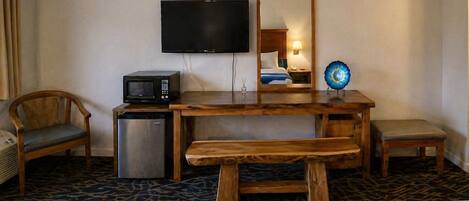 A hotel room with a wooden desk, a television, a microwave, a small refrigerator, a bench, and a bed with a blue bedspread.