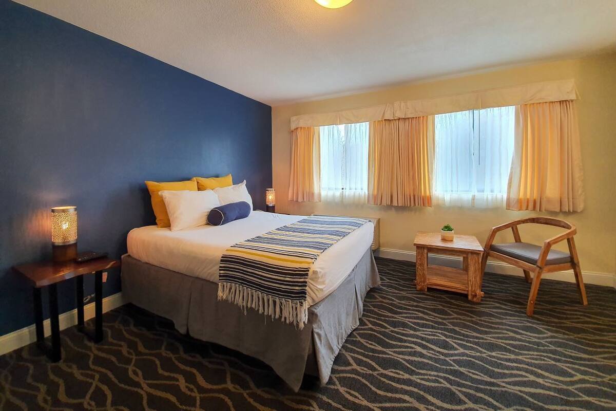 Una habitación de hotel con cama, mesita de noche, lámpara, silla y ventanas con cortinas.