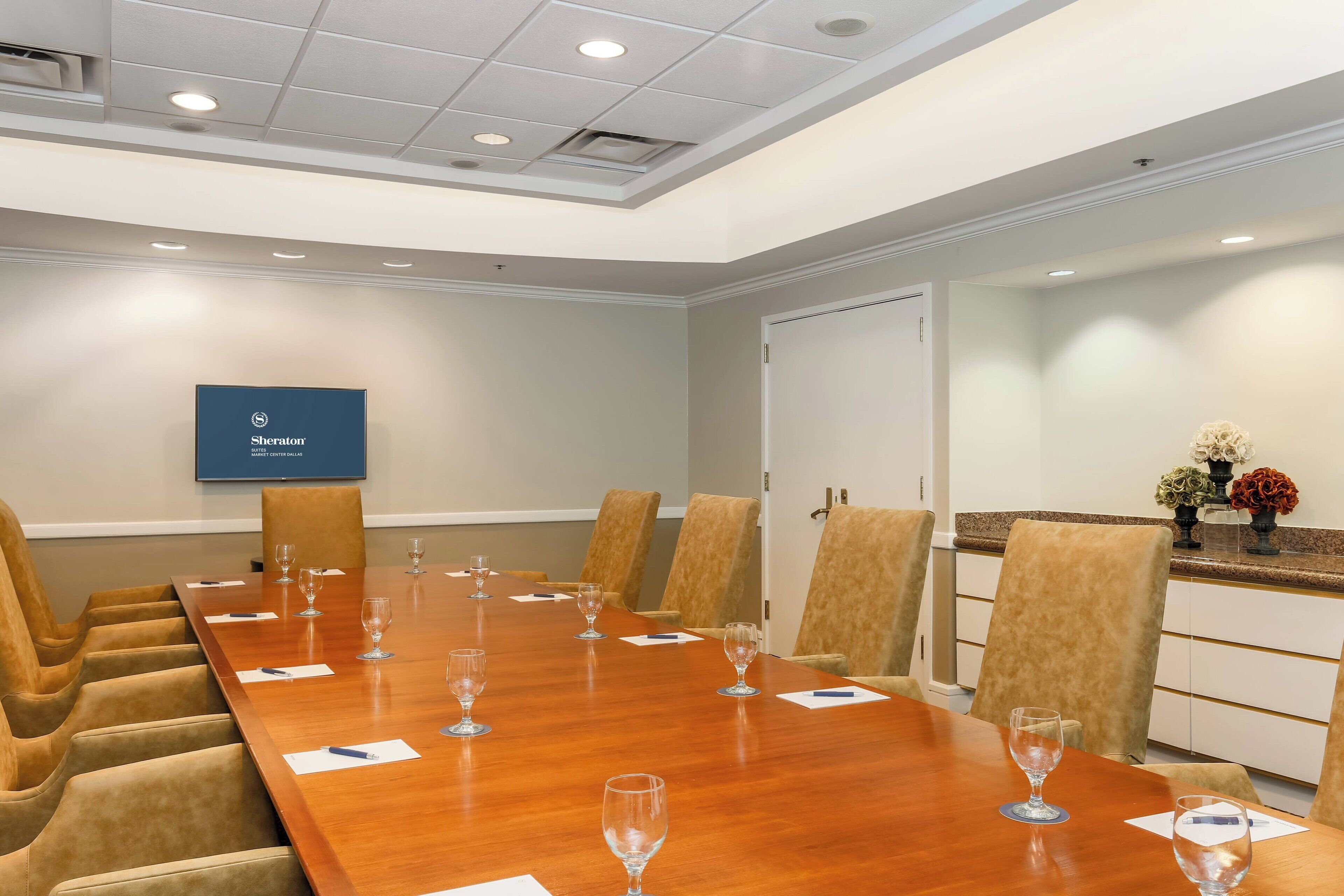 A conference room with a long wooden table, beige chairs, and a flat-screen TV on the wall.