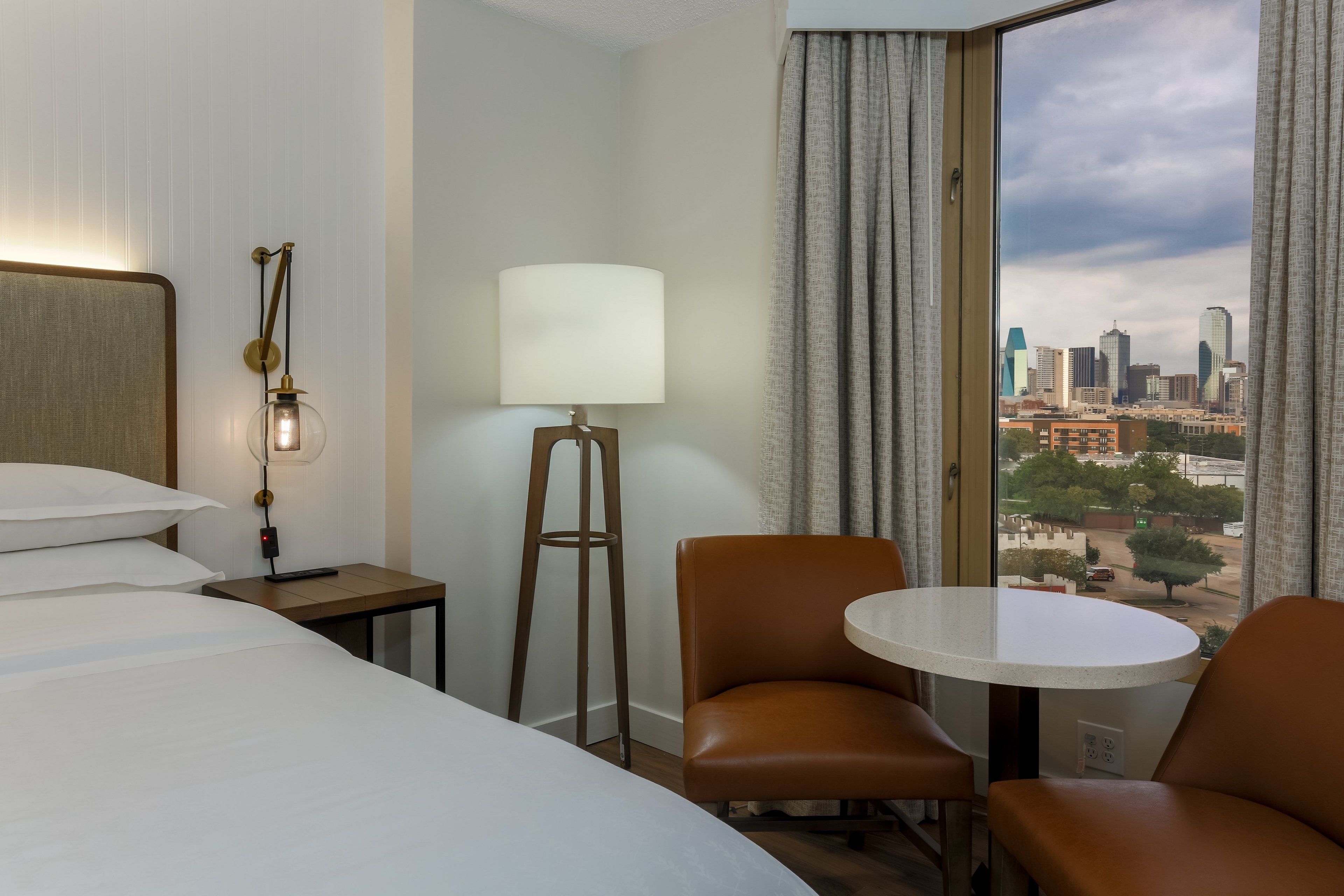 Une chambre d’hôtel avec un lit, un bureau, une lampe, une chaise et une vue sur le paysage urbain par la fenêtre.