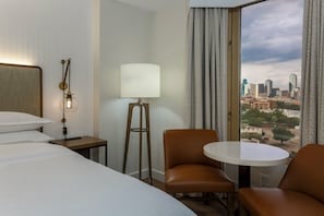 A hotel room with a bed, a desk, a lamp, a chair, and a view of a cityscape through a window. - Sheraton Suites Market Center Dallas (Dallas)