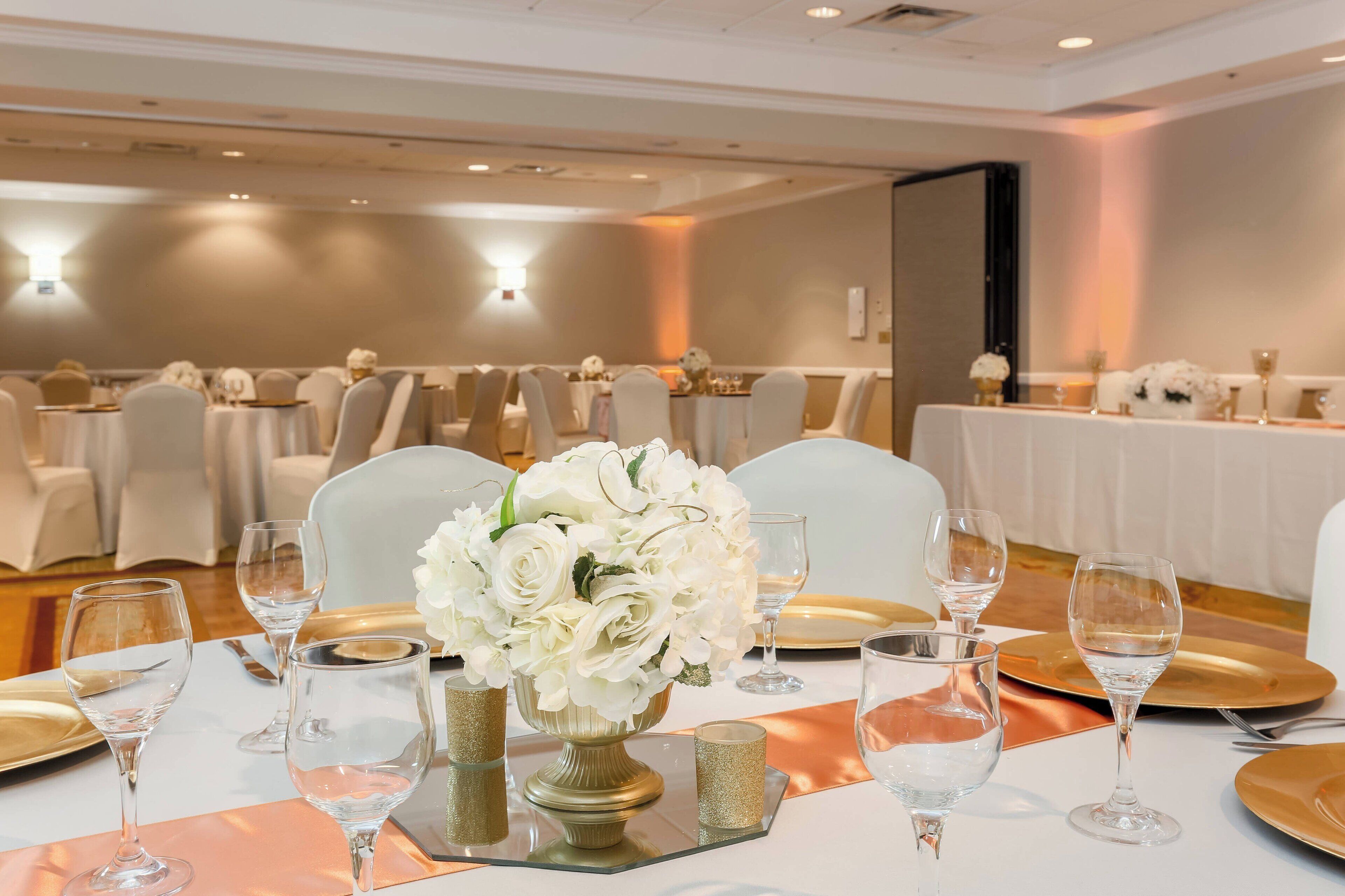 Un salón de banquetes con mesas redondas dispuestas para un evento formal, con centros de mesa florales blancos y vajilla dorada.