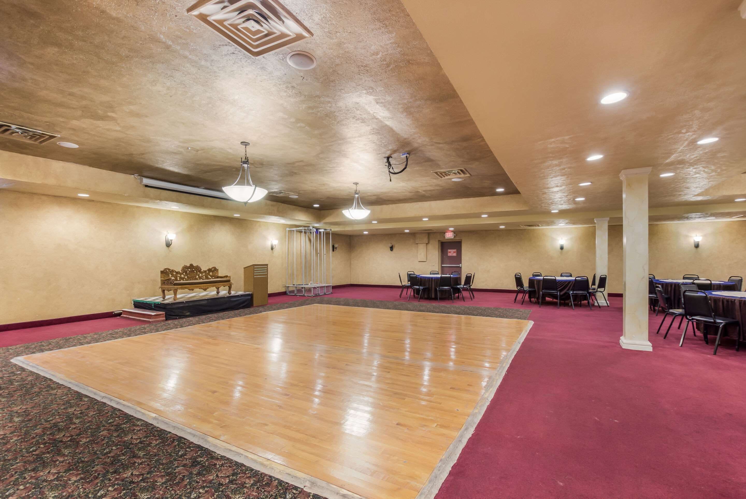 A spacious room with a large wooden dance floor, multiple tables with chairs, and a red carpeted area.