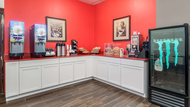 Een keukenhoek met rode wanden, witte kastjes en een koelkast. Er staan sapdispensers, een koffieapparaat en diverse etenswaren op het aanrecht.