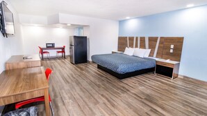 Een hotelkamer met een houten vloer, een bed met een blauw beddegoed met patroon, een bureau met stoel, een koelkast en een prullenbak.