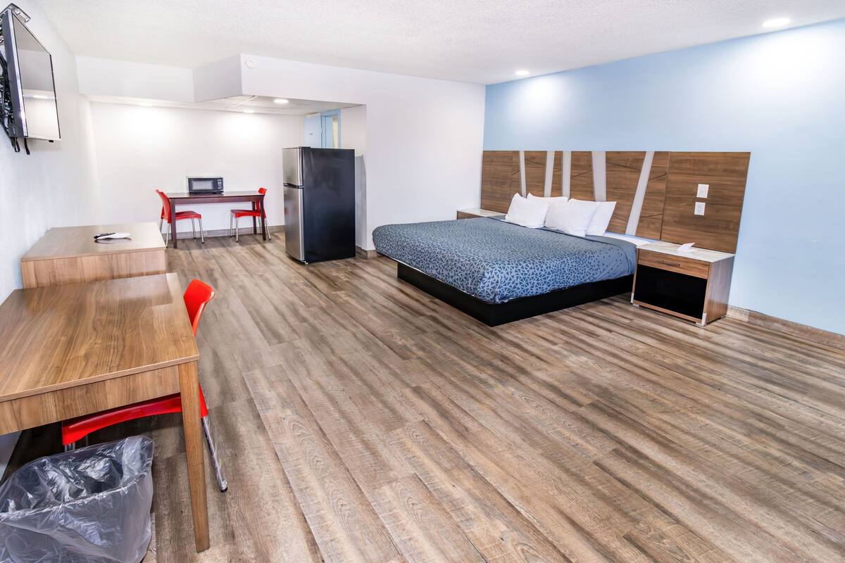 Habitación de hotel con piso de madera, cama con colcha azul estampada, escritorio con silla, refrigerador y un cubo de basura.
