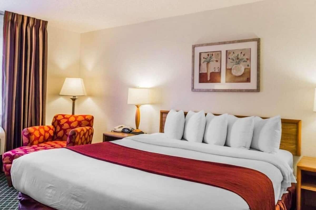 Una camera d'albergo con un letto, due lampade ai comodini, una sedia, un tavolino e un quadro appeso al muro.
