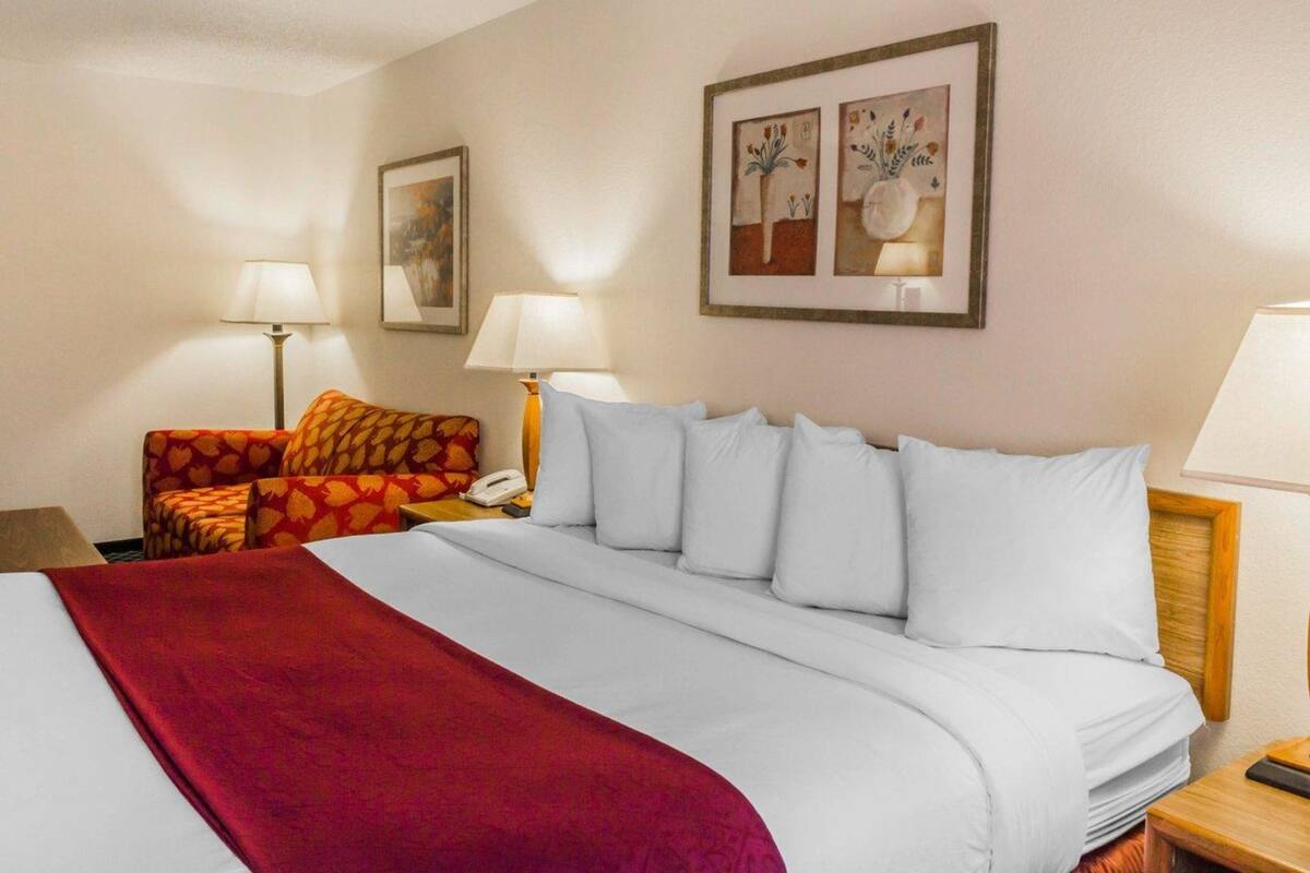 Una camera d'albergo con un letto grande, una coperta rossa, una poltrona con motivi e due lampade ai comodini, oltre a quadri appesi alle pareti.