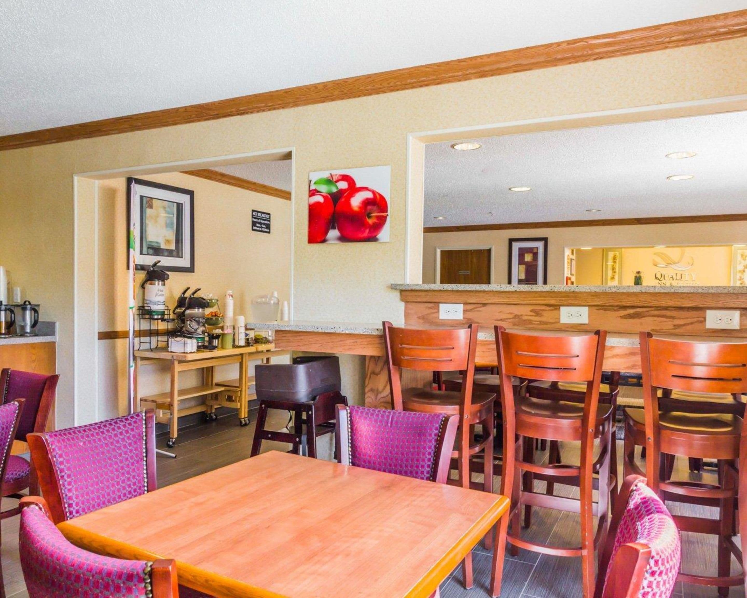 Un comedor con mesas y sillas de madera, una zona de cocina con cafetera y una pared decorada con un cuadro de frutas.