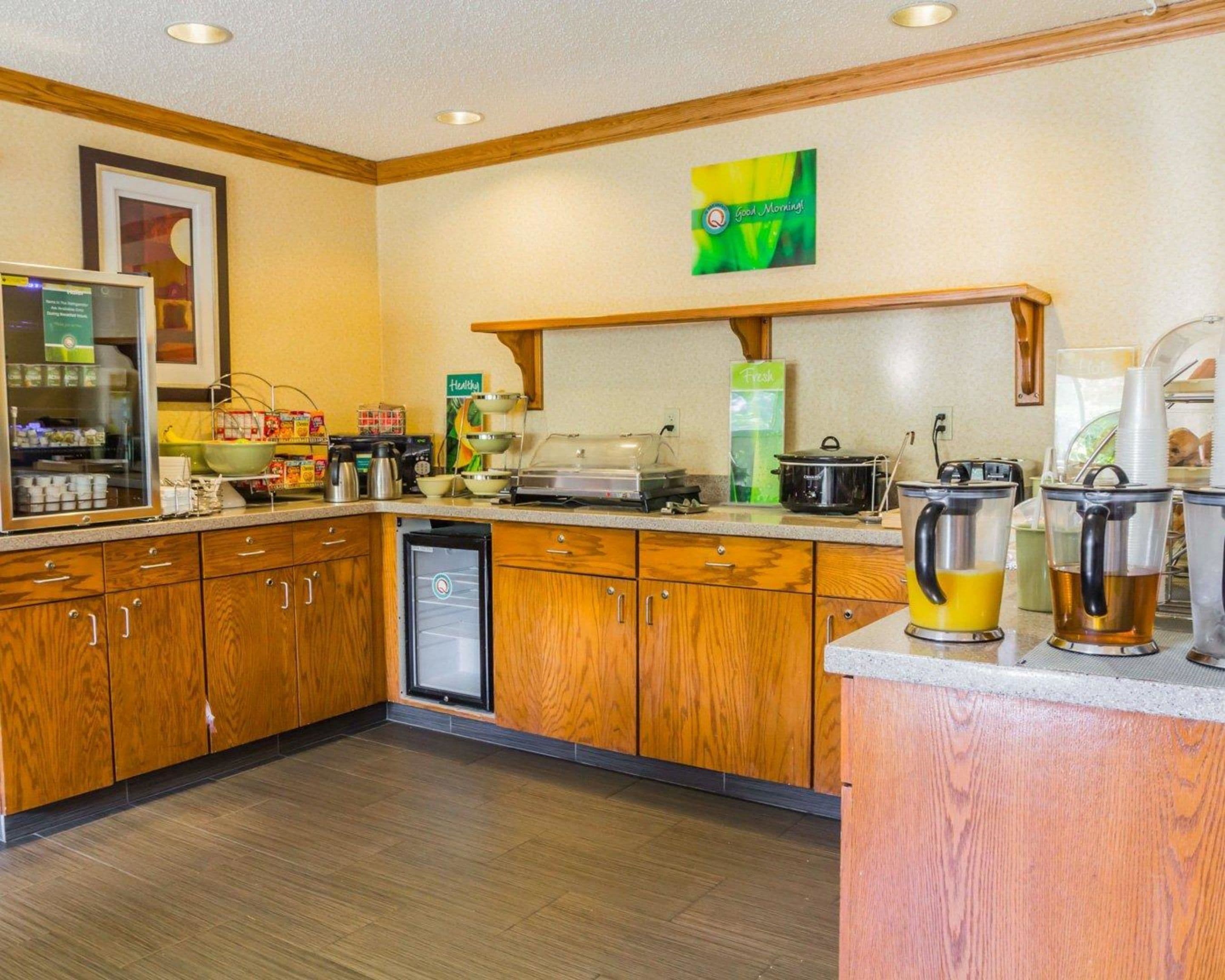 A hotel kitchen with wooden cabinets, a coffee station, a refrigerator, and a breakfast bar.