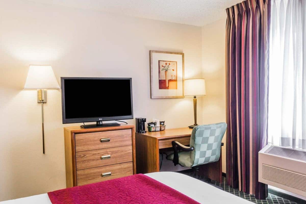 Una camera d'albergo con un letto, una scrivania, una sedia, una televisione, una lampada, un portafoto e una finestra con tende.