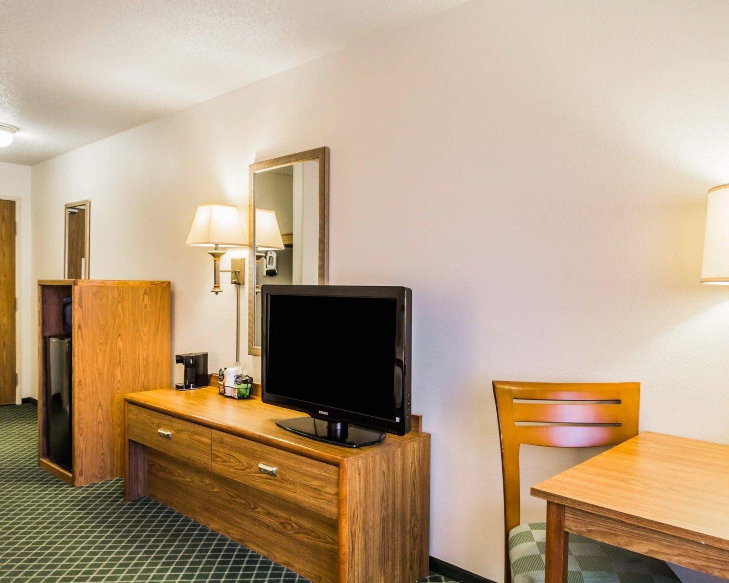 A hotel room with a wooden dresser, a television, a lamp, a mirror, and a chair.