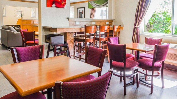 A hotel dining area with wooden tables and chairs, a counter with a green backdrop, and a view of greenery outside. - Quality Inn & Suites Golden - Denver West (Golden)