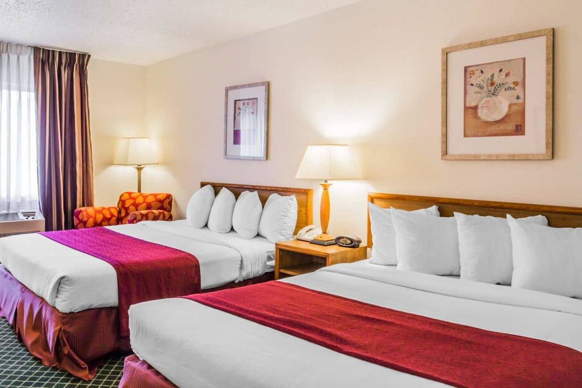 Camera d'albergo con due letti, ciascuno con lenzuola bianche e coperte rosse. Due comodini con lampade, un quadro incorniciato al muro e una finestra con tende.