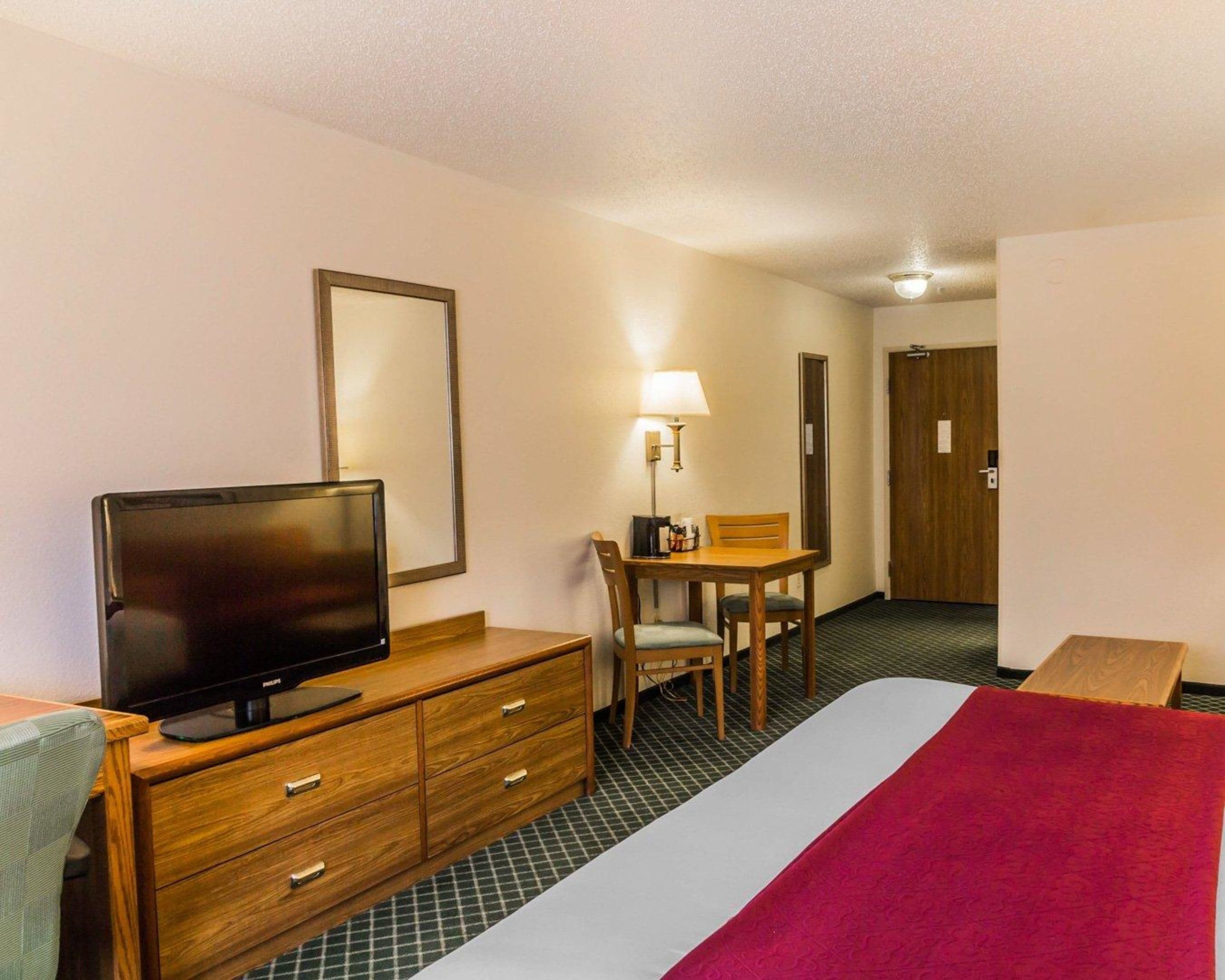 A hotel room with a bed, a television on a wooden dresser, a desk with a chair, and a lamp.