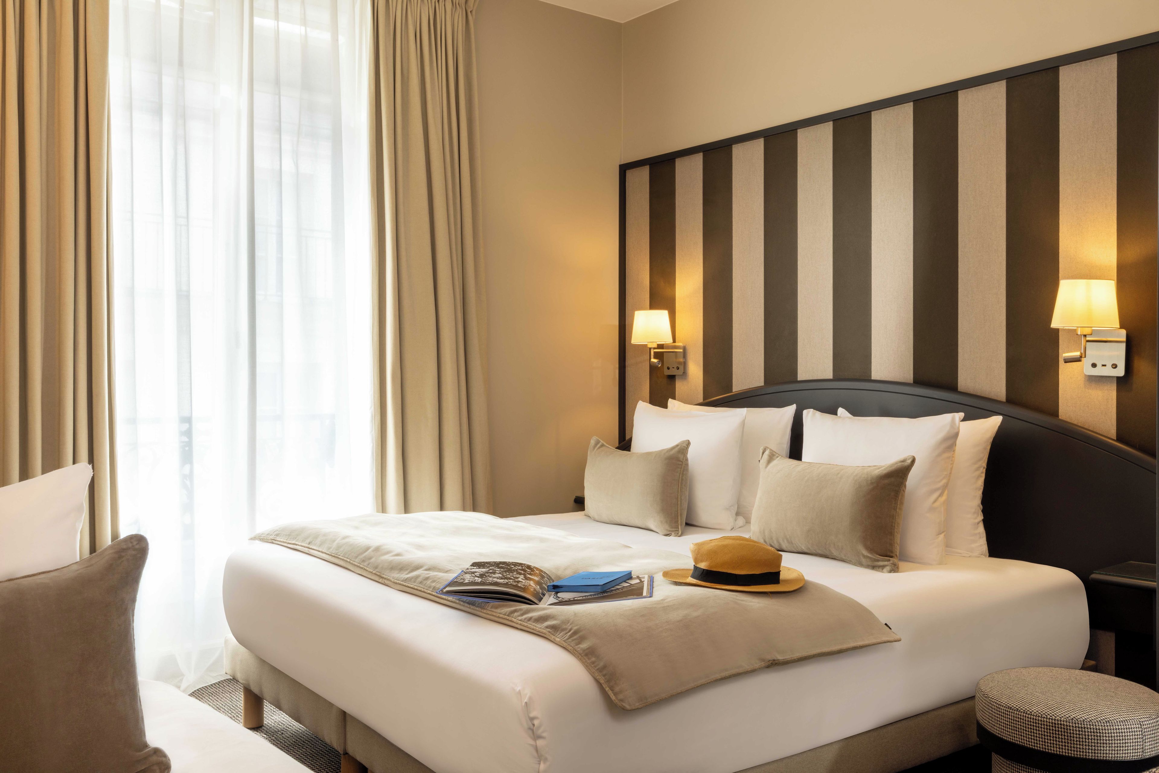 A hotel room with a bed, pillows, a nightstand, a lamp, and a window with curtains.