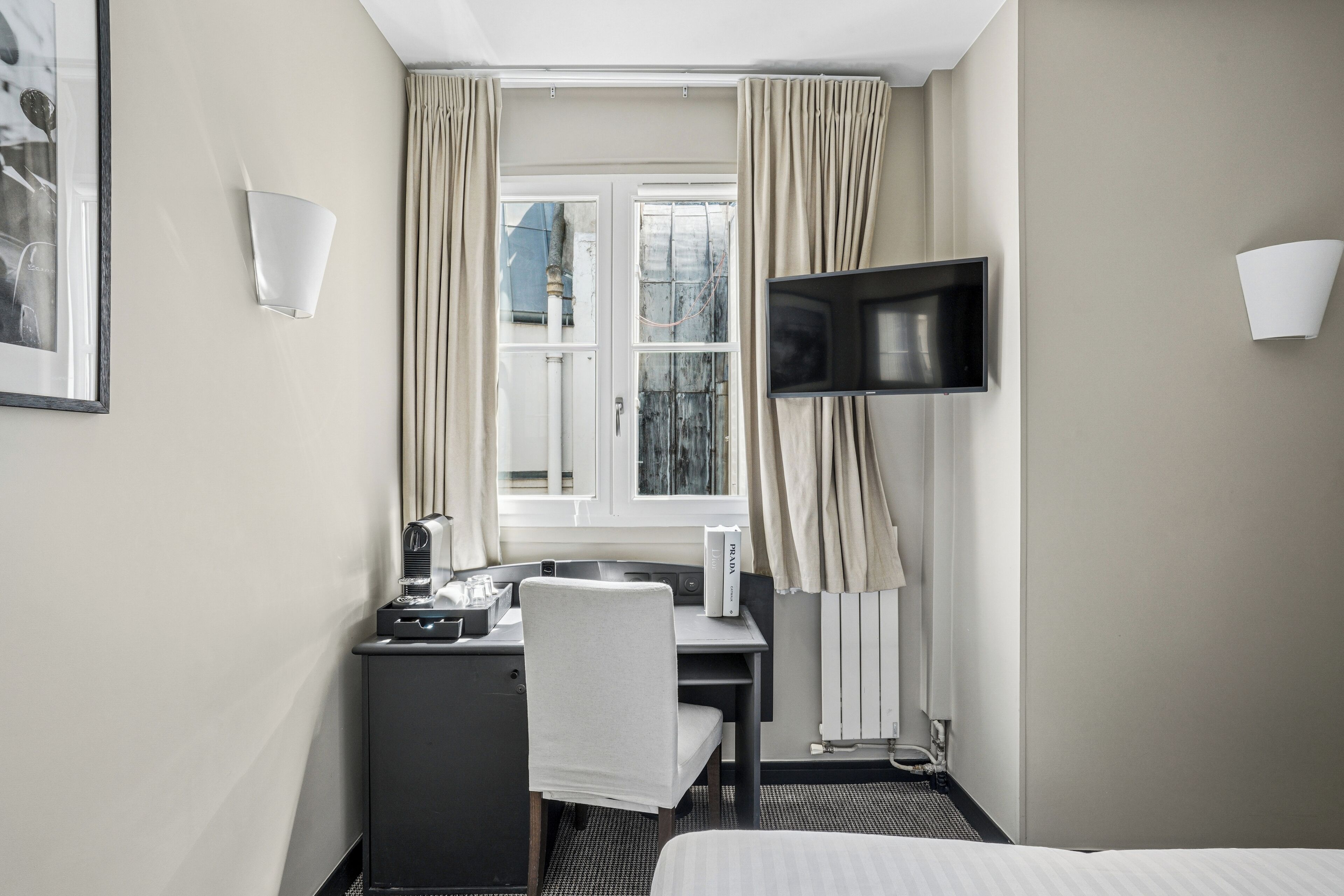 A hotel room with a bed, a desk with a chair, a television mounted on the wall, and a window with curtains.