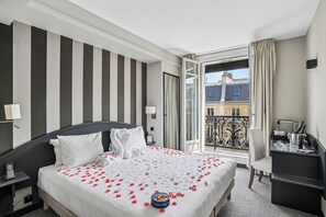 A hotel room with a large bed, a bedside table with a phone, a chair, a desk with a lamp, and a balcony with a view of buildings.