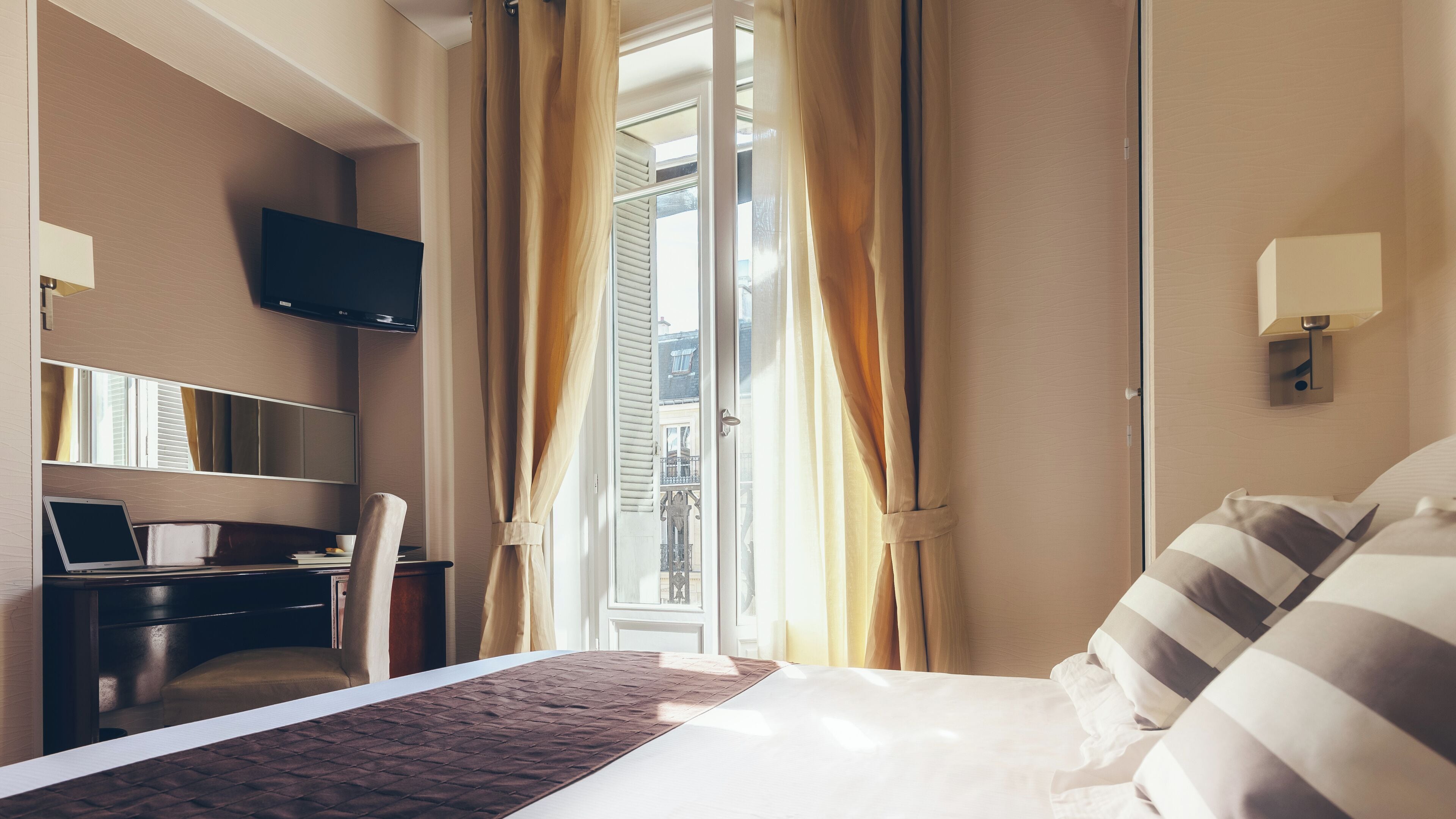 Habitación de hotel con cama, escritorio con una computadora portátil, televisor montado en la pared y ventana con cortinas.