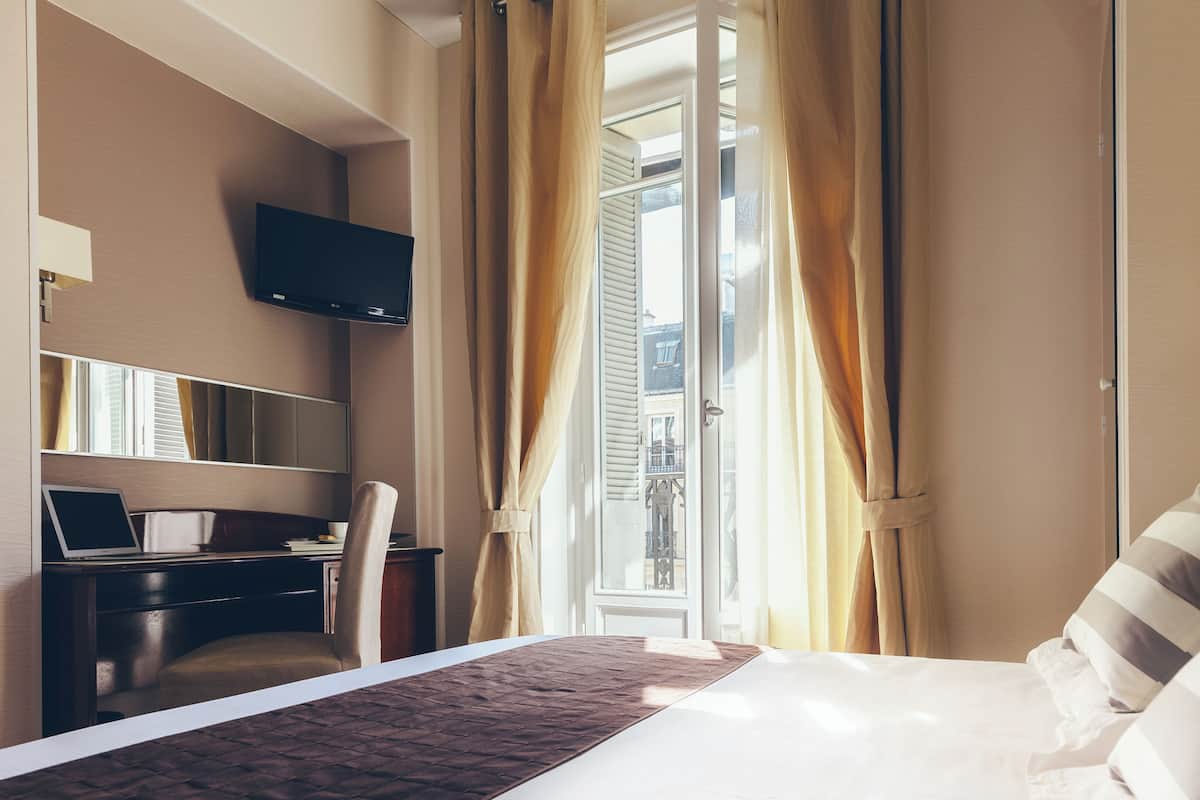 A hotel room with a bed, a desk with a laptop, a TV mounted on the wall, and a window with curtains.