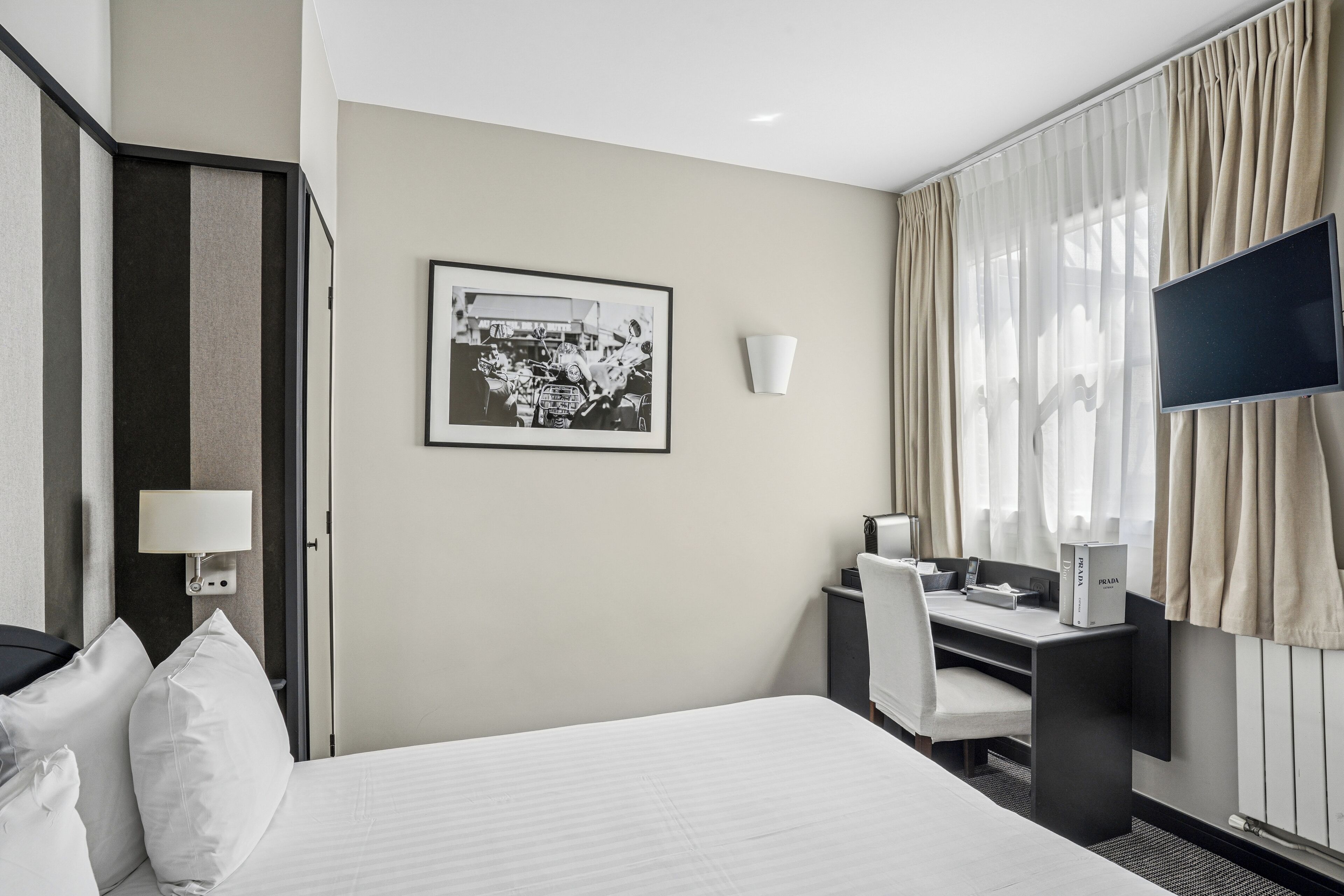 Une chambre d’hôtel avec un lit, un bureau et une chaise, une télévision, un tableau encadré au mur et une fenêtre avec des rideaux.