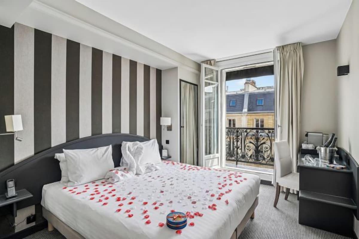 A hotel room with a bed, a desk, a chair, a balcony, and a view of buildings.