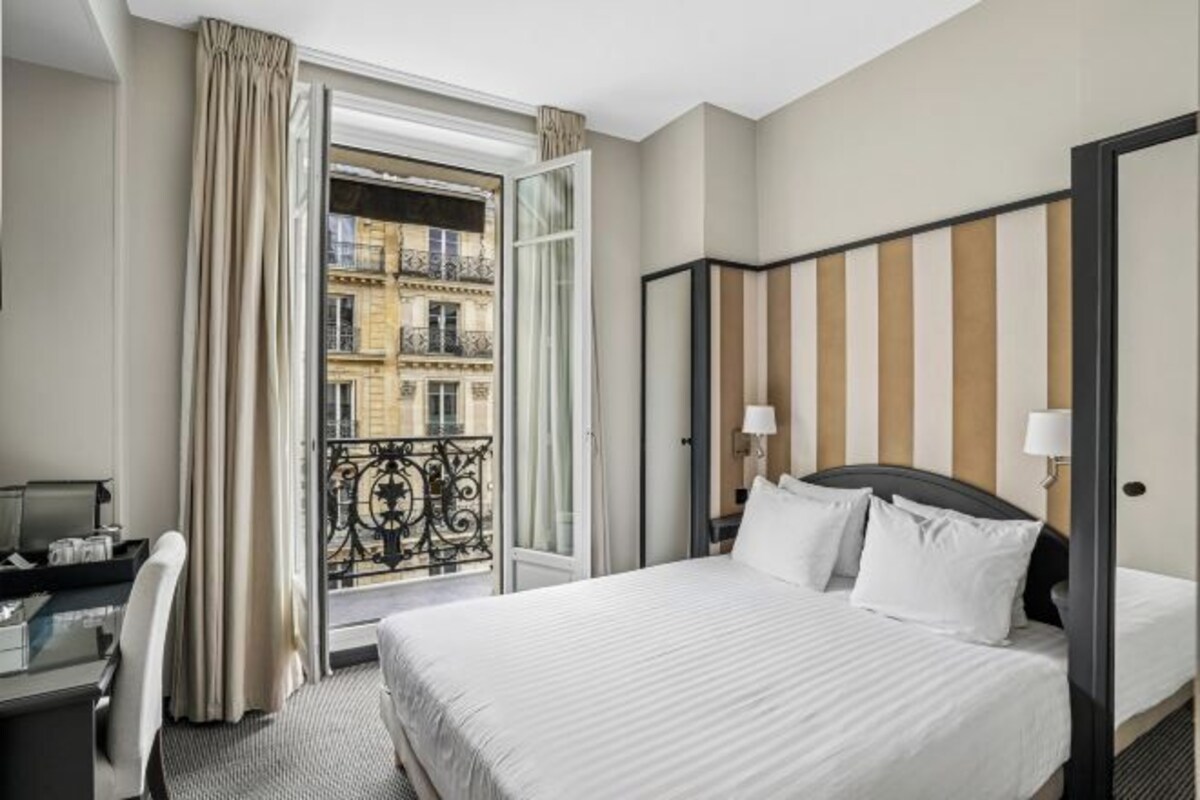 A hotel room with a bed, a desk, a chair, a window with curtains, and a view of a building.