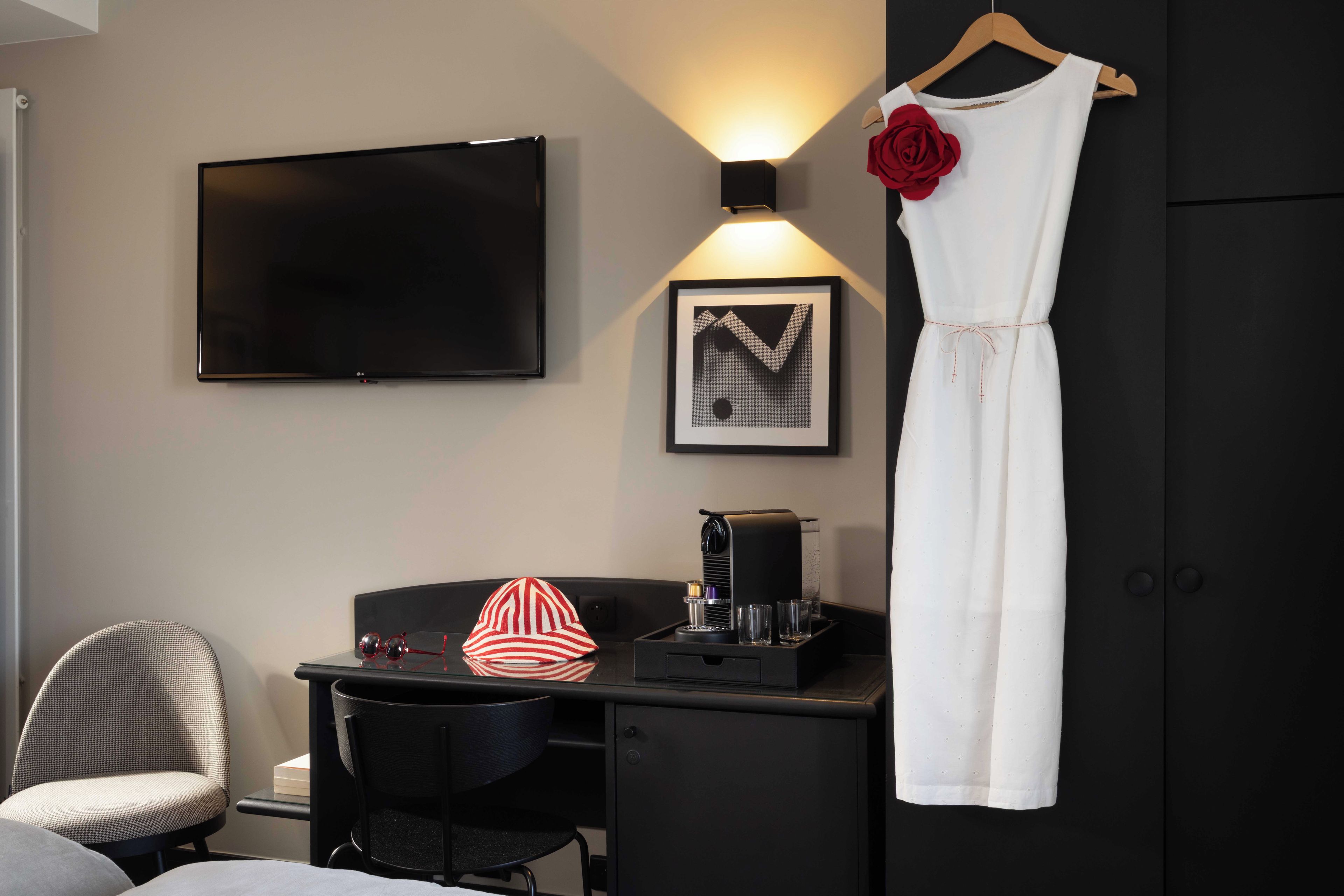 Habitación de hotel con televisión de pantalla plana, un cuadro enmarcado, máquina de café, escritorio, silla y un vestido colgado.