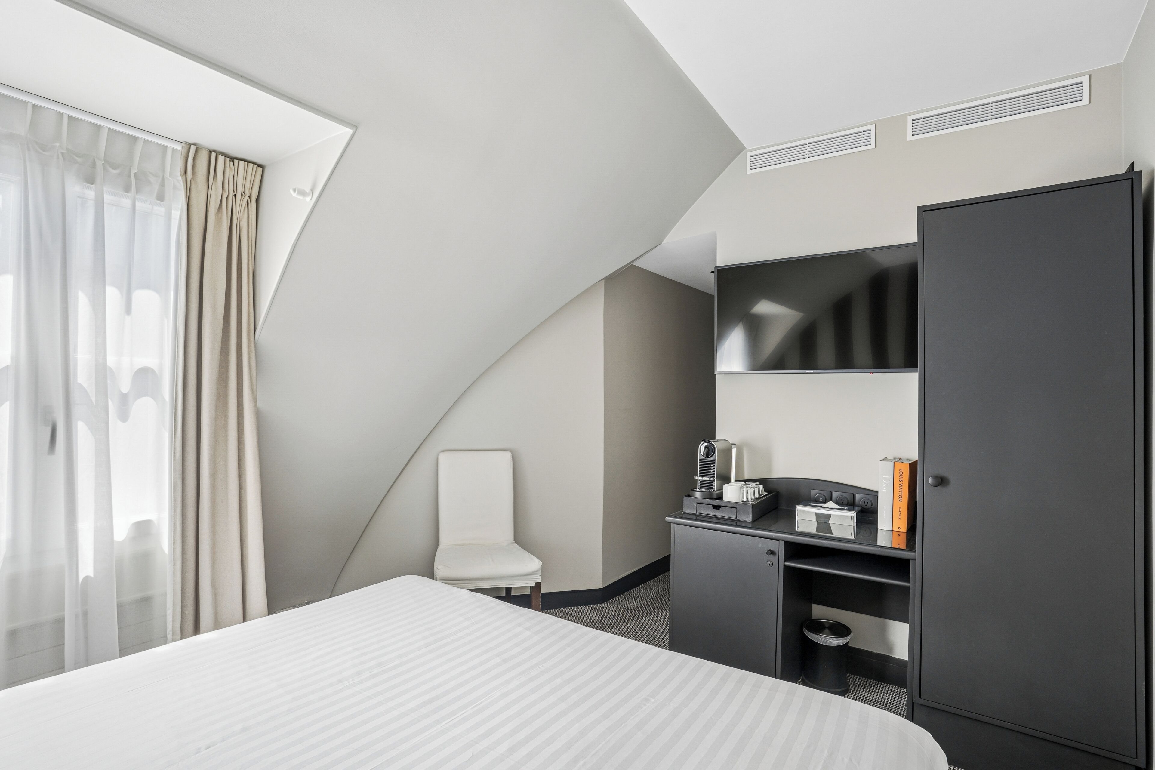 A hotel room with a bed, a desk, a chair, a wardrobe, a television, and a window with curtains.