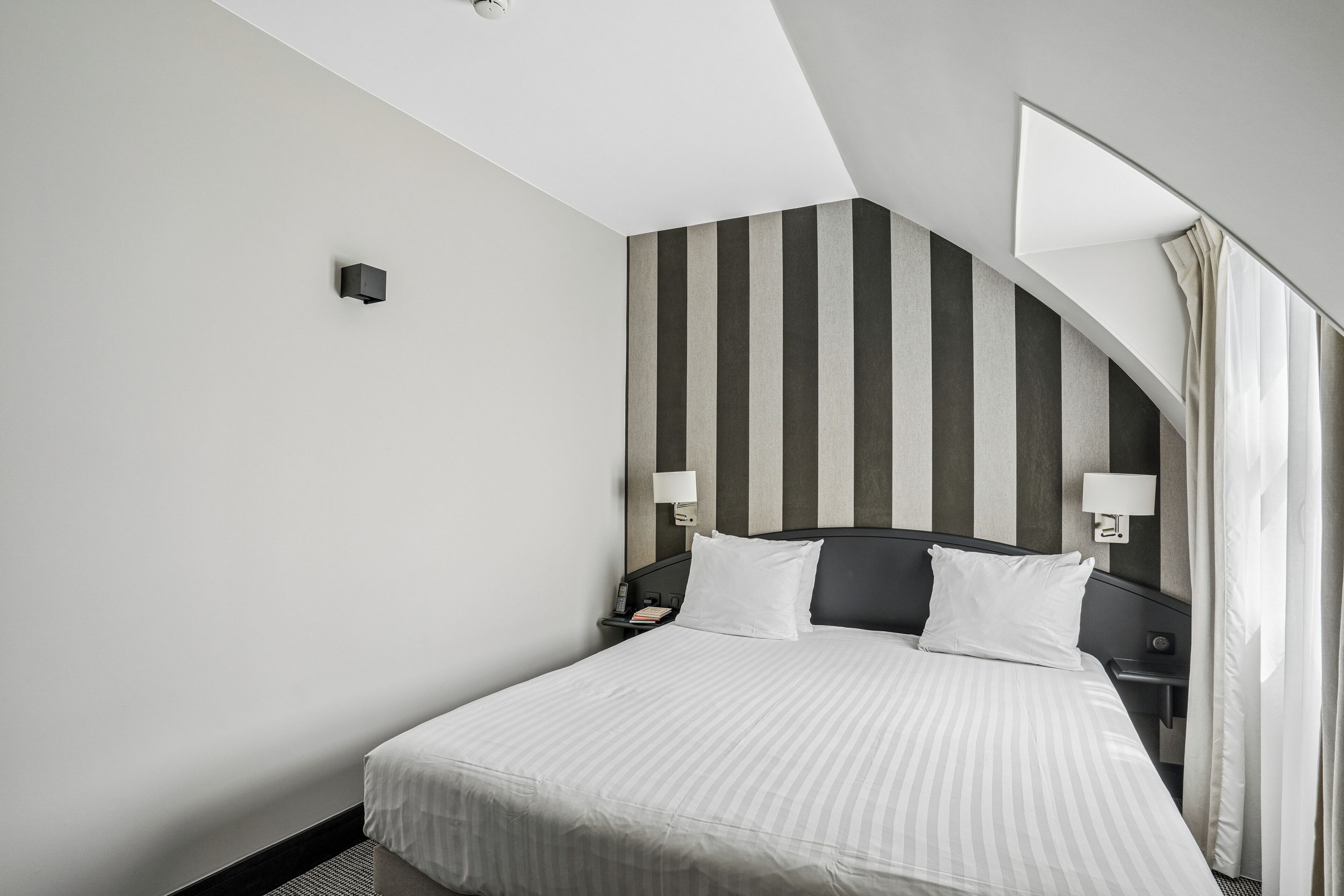Une chambre d’hôtel comprenant un lit, deux tables de chevet, une petite table, une lampe fixée au mur et un grand miroir.