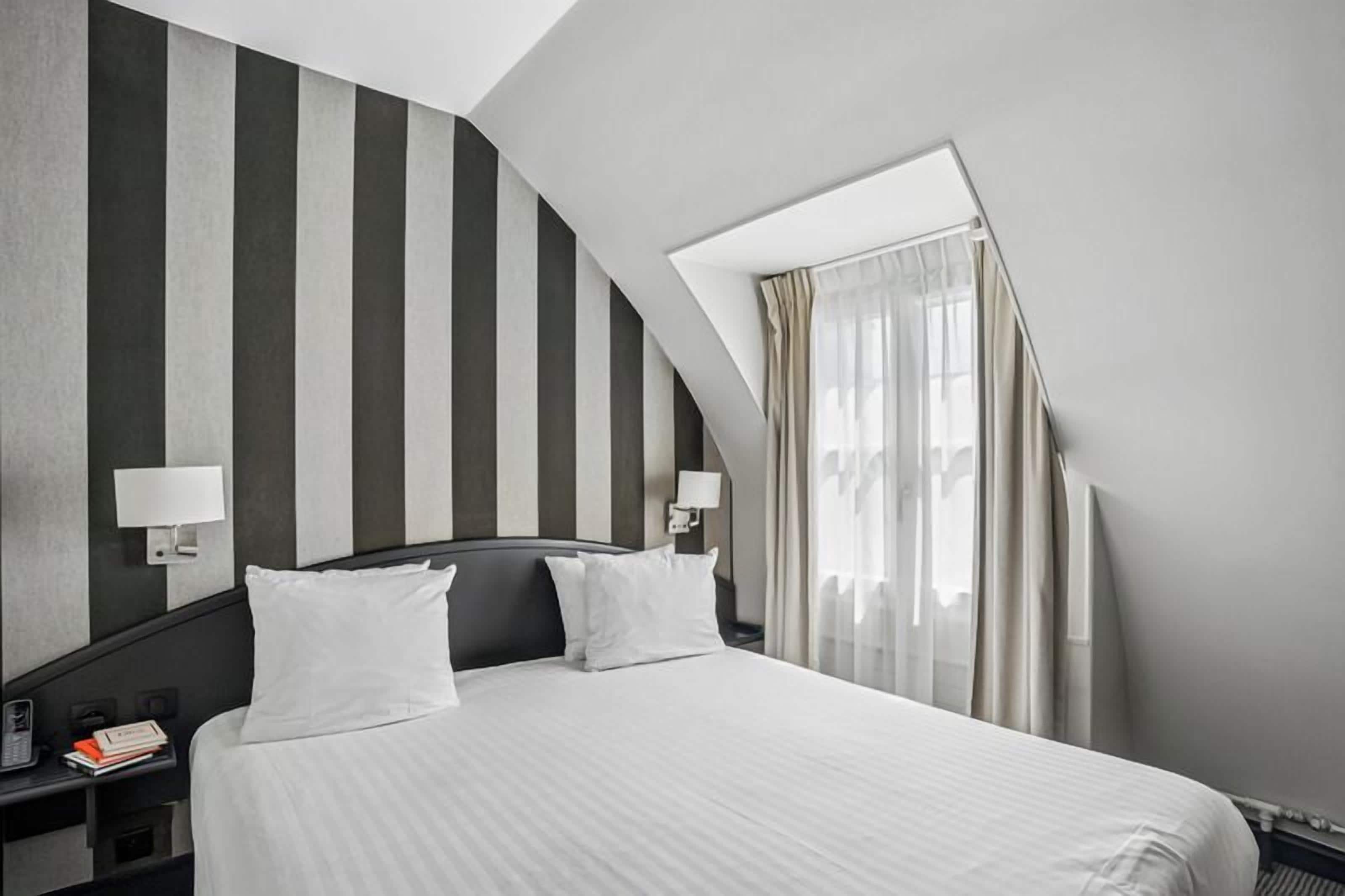 Una habitación de hotel con cama, dos mesitas de noche, una lámpara de noche, una ventana con cortinas y un techo inclinado.