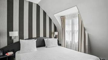 Una habitación de hotel con cama, dos mesitas de noche, una lámpara de noche, una ventana con cortinas y un techo inclinado.
