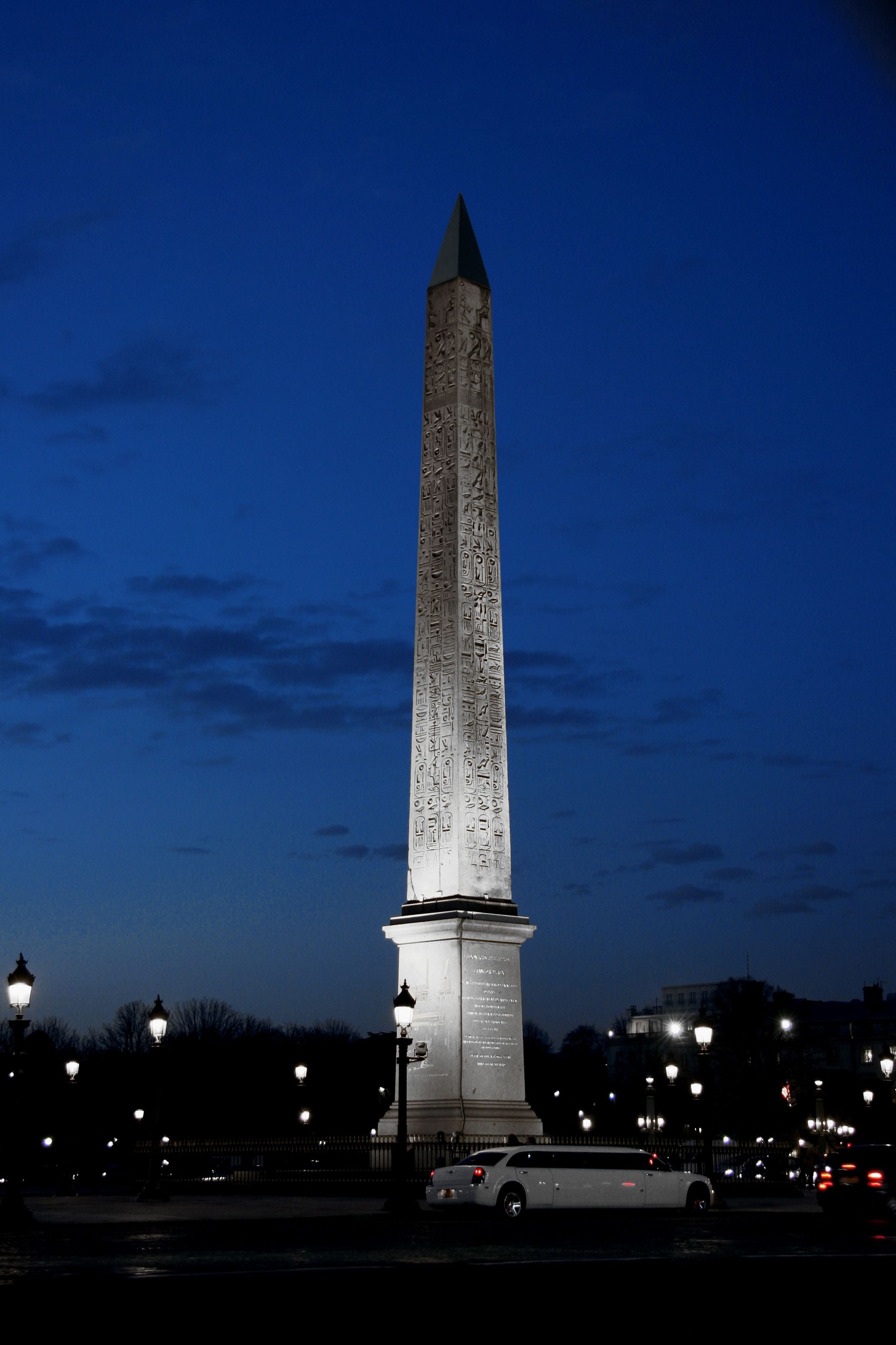 A tall, narrow monument with hieroglyphs, surrounded by street lamps and a parked car.